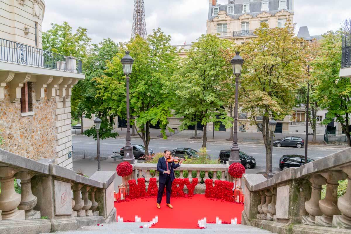 Paris proposal at Rue de Camoëns with Eiffel Tower view, red carpet, 'Marry Me' letters, and a master violinist performing in a romantic Parisian street setting