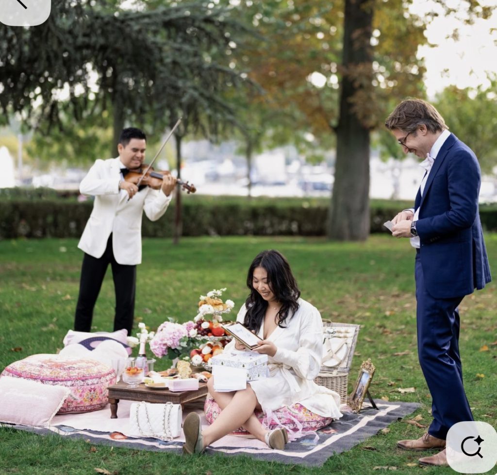 Woman sitting on a picnic blanket in Paris opening a love letter as a violinist plays during a surprise proposal