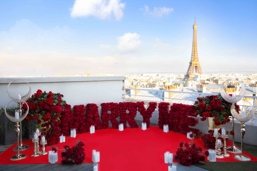 Elegant Paris rooftop proposal with red rose “Marry Me” letters, white carpet, gold candle holders, and panoramic Eiffel Tower view under a clear blue sky.