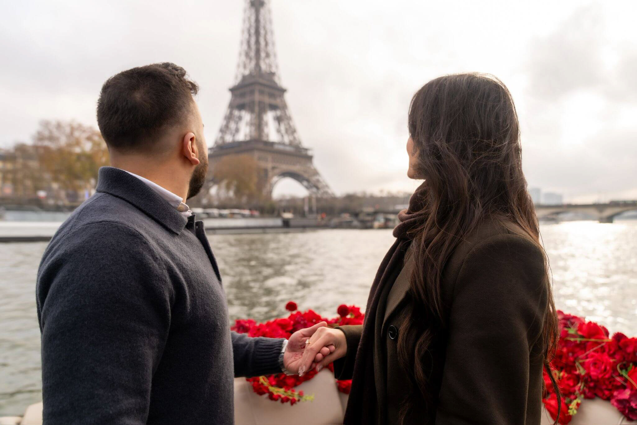 A couple standing together on a private boat in Paris, surrounded by red rose decorations while cruising on the Seine River. The Eiffel Tower is visible in the background, creating a romantic setting after a boat proposal in Paris. This private Seine River proposal offers Eiffel Tower views, an intimate atmosphere, and a refined experience for couples planning a proposal in Paris.