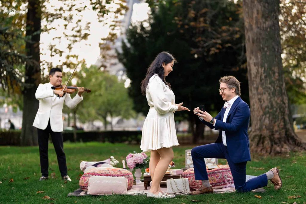 Romantic picnic proposal in Paris with Eiffel Tower view, violinist playing live music as man proposes on one knee