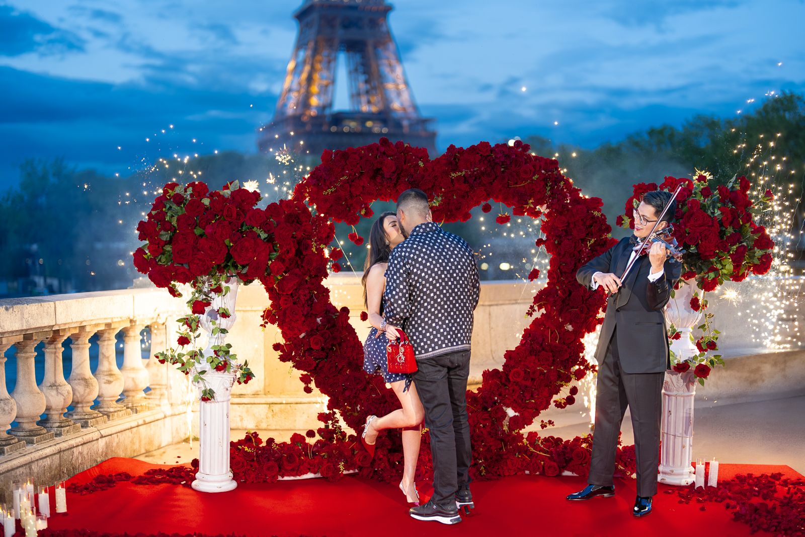 Paris proposal at Bir-Hakeim bridge during blue hour with a Big Love Heart red rose arch, live violinist, sparklers, red carpet, and candles, with the Eiffel Tower in the background — setup and coordination by Events in Paris