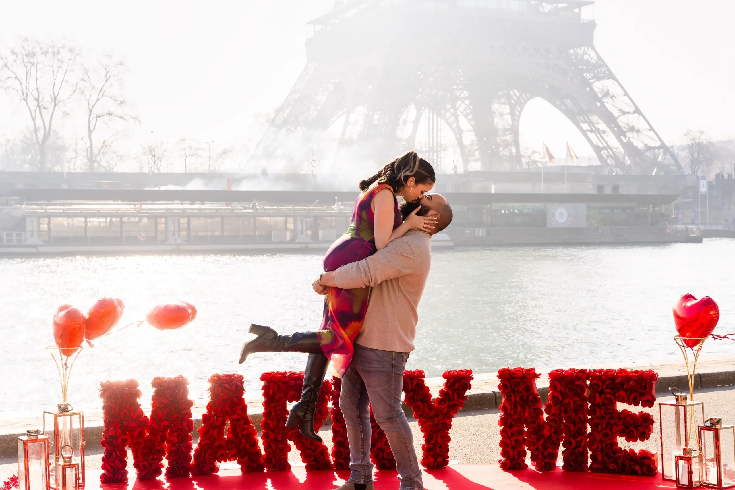 Couple celebrating a marriage proposal on the Seine riverbank in Paris, with red rose "Marry Me" letters, heart balloons, and the Eiffel Tower in the background