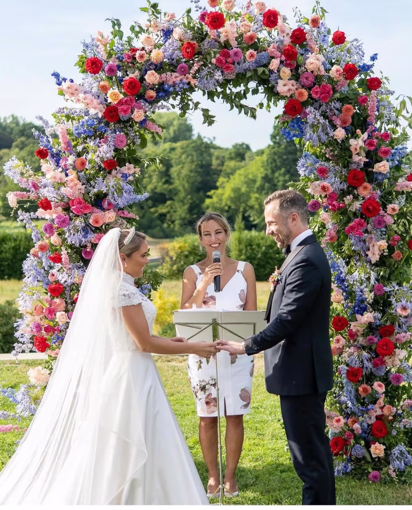Aude Abadie officiating an outdoor wedding ceremony under a large colorful floral arch with red, pink and blue blooms, bride in white gown and groom in navy tuxedo holding hands beside her