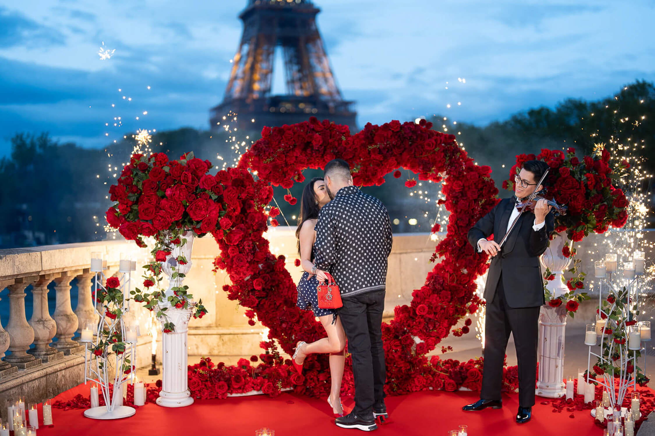 Couple inside a red rose Big Love Heart arch on a red carpet at Bir-Hakeim, with a live violinist, firework fountains, and the Eiffel Tower lit up at blue hour