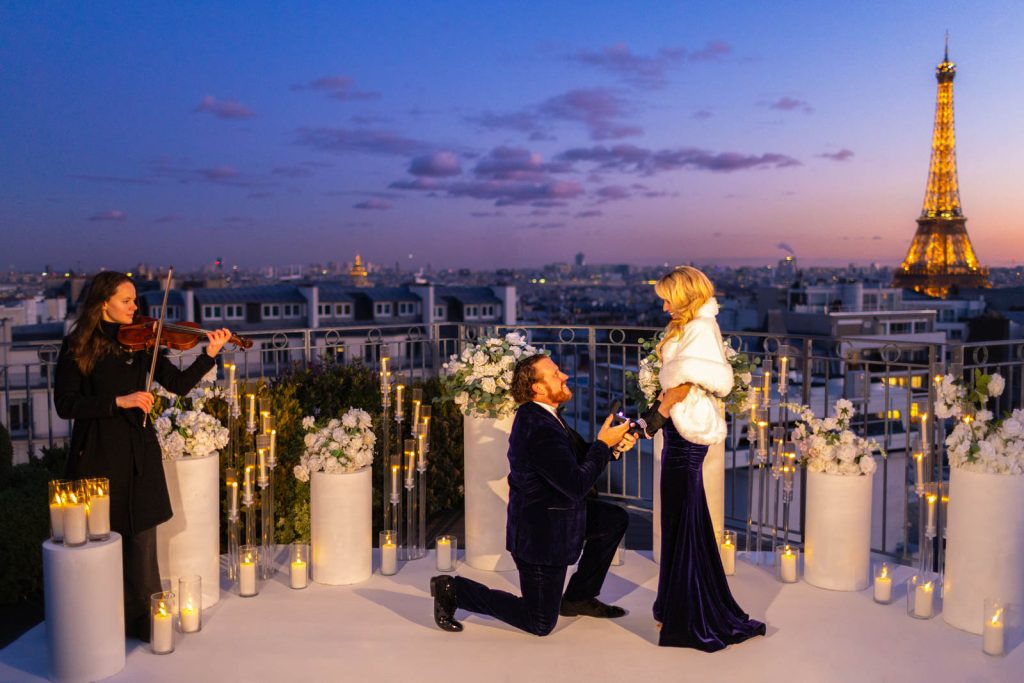 Man on one knee proposing on the Copernic rooftop in Paris at blue hour, white floral columns, candles and transparent candelabras surrounding the couple, violinist playing, illuminated Eiffel Tower behind