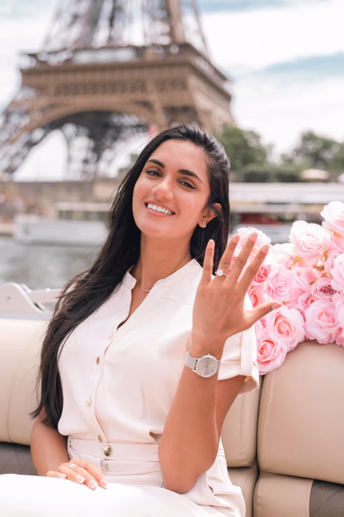 Woman smiling and showing her engagement ring on a private boat on the Seine, pink roses beside her, Eiffel Tower directly behind