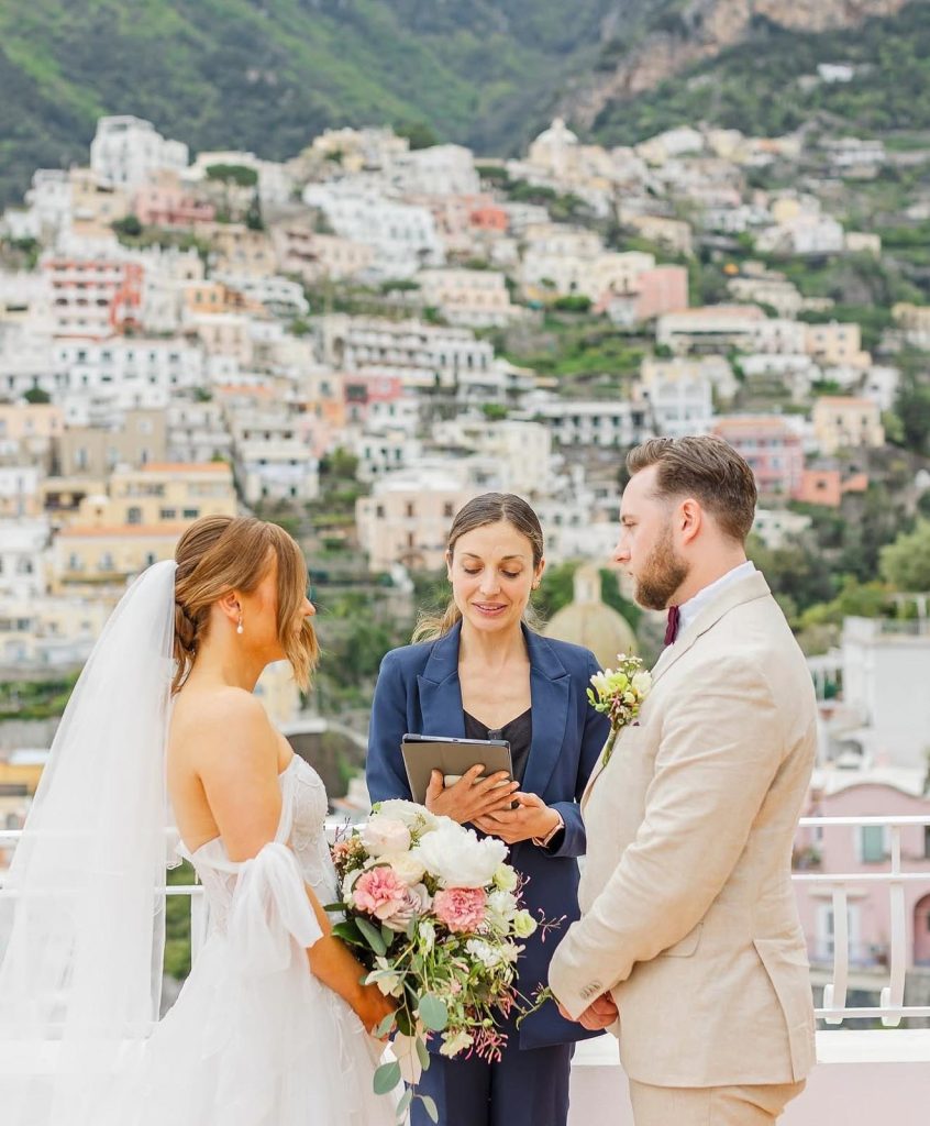 Love Gracefully officiant reading from tablet during elopement ceremony on a terrace in Positano, bride in off-shoulder lace gown with pink and white bouquet and groom in beige suit, colorful Italian hillside village behind