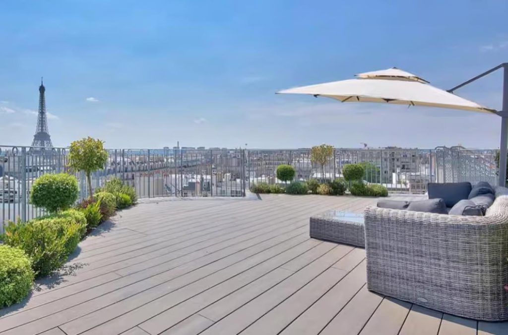 Large private rooftop terrace in Paris with wood decking, rattan sofa, parasol, boxwood topiaries and Eiffel Tower visible on the left across the Paris skyline under a clear blue sky