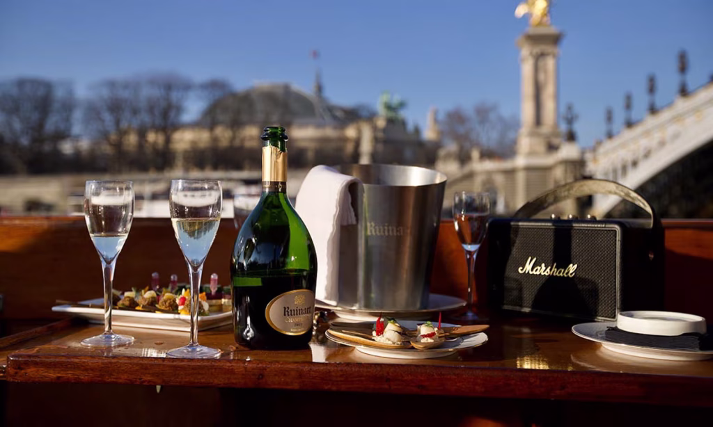 Ruinart champagne bottle in silver ice bucket with two crystal flutes, canapés and Marshall speaker on the wooden deck of a private yacht on the Seine river in Paris, Pont Alexandre III and Grand Palais softly blurred behind