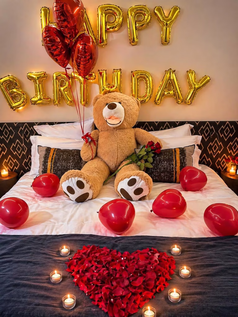 Paris hotel room birthday decoration, large teddy bear holding red roses on a white bed surrounded by red heart balloons, gold Happy Birthday foil balloon letters on the wall, red rose petal heart with candles on the floor, candles on both bedside tables