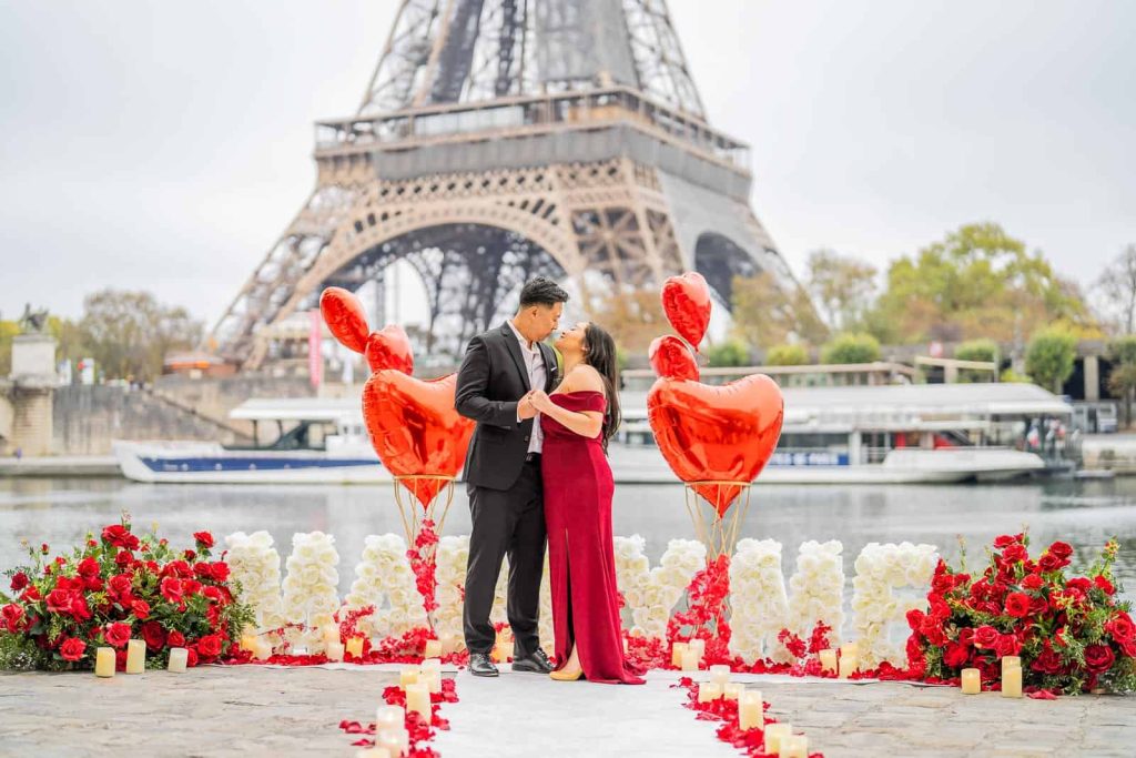 Couple kissing during a Paris proposal on the Seine riverbank, man in black suit, woman in burgundy off-shoulder gown, white rose Marry Me letter display, red rose arrangements, heart-shaped red balloons, candles and rose petals on the ground, Eiffel Tower centered directly behind