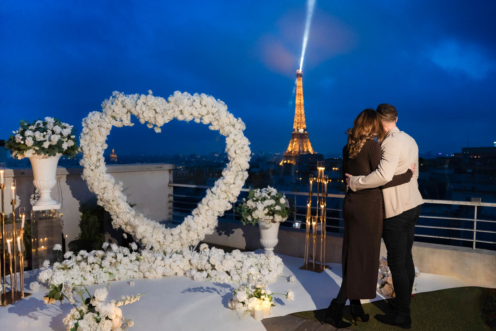 Couple embracing after proposal on the Sablon rooftop terrace in Paris at blue hour, white heart-shaped floral arch beside them, illuminated Eiffel Tower with light beam behind