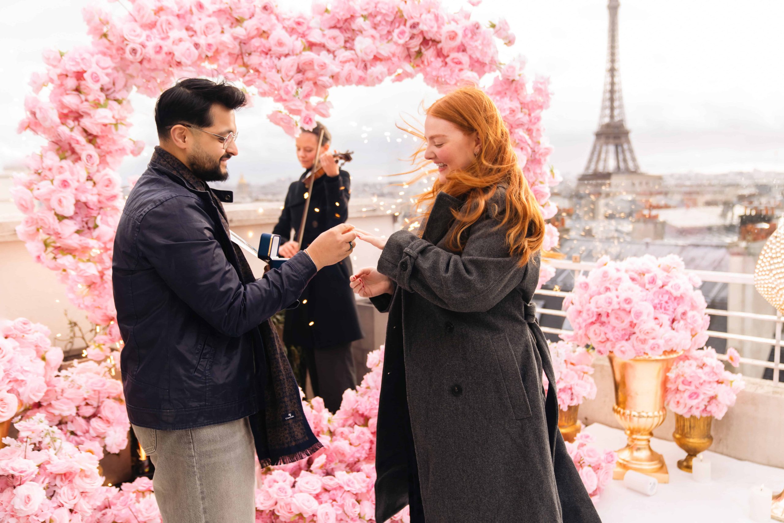 Man sliding engagement ring onto fiancée's finger inside a pink rose heart arch on the Sablon rooftop in Paris, violinist playing behind them, sparklers lit, Eiffel Tower visible behind