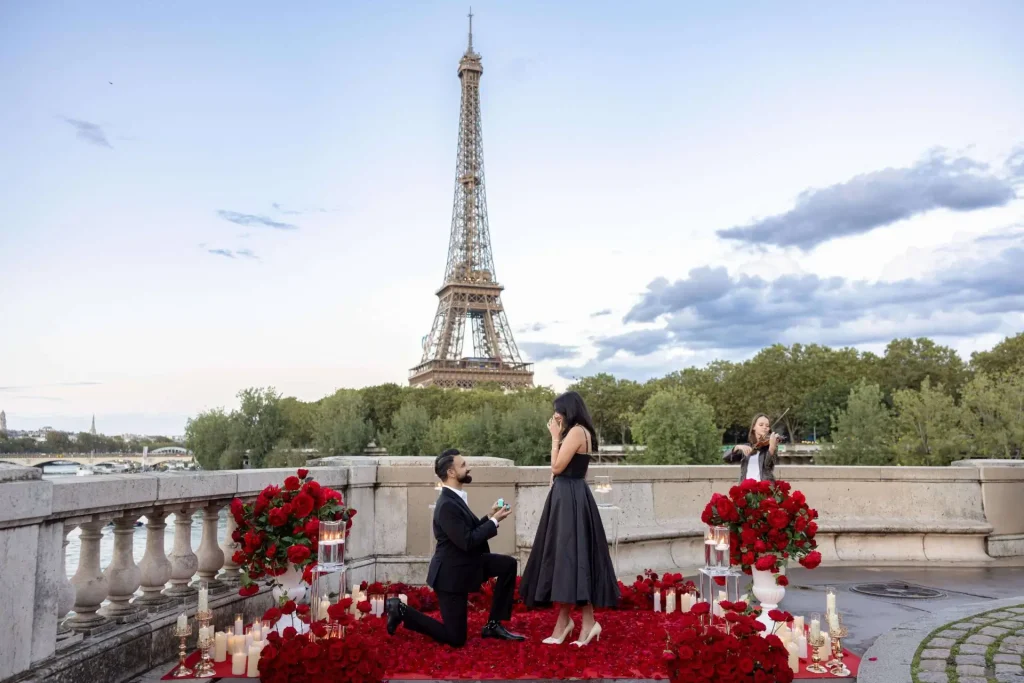 Heiratsantrag vor dem Eiffelturm an der Bir-Hakeim-Brücke mit einem Mann auf einem Knie, roten Rosen, Kerzen und einem Live-Geiger