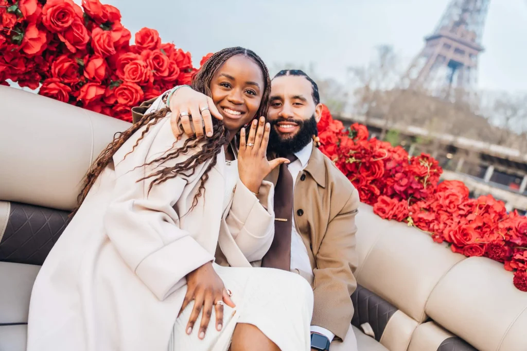 Casal recém-noivo exibindo o anel de noivado em um barco no Sena, com rosas vermelhas e a Torre Eiffel ao fundo, após o pedido de casamento em Paris