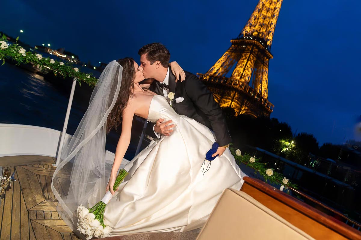 Paris elopement on private champagne yacht with bride in white ball gown and groom kissing, Eiffel Tower at night