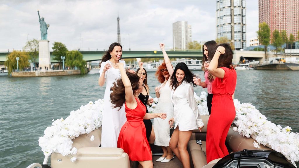 Bachelorette party group dancing on flower-decorated private boat on the Seine with Eiffel Tower and Paris Statue of Liberty in background, Paris