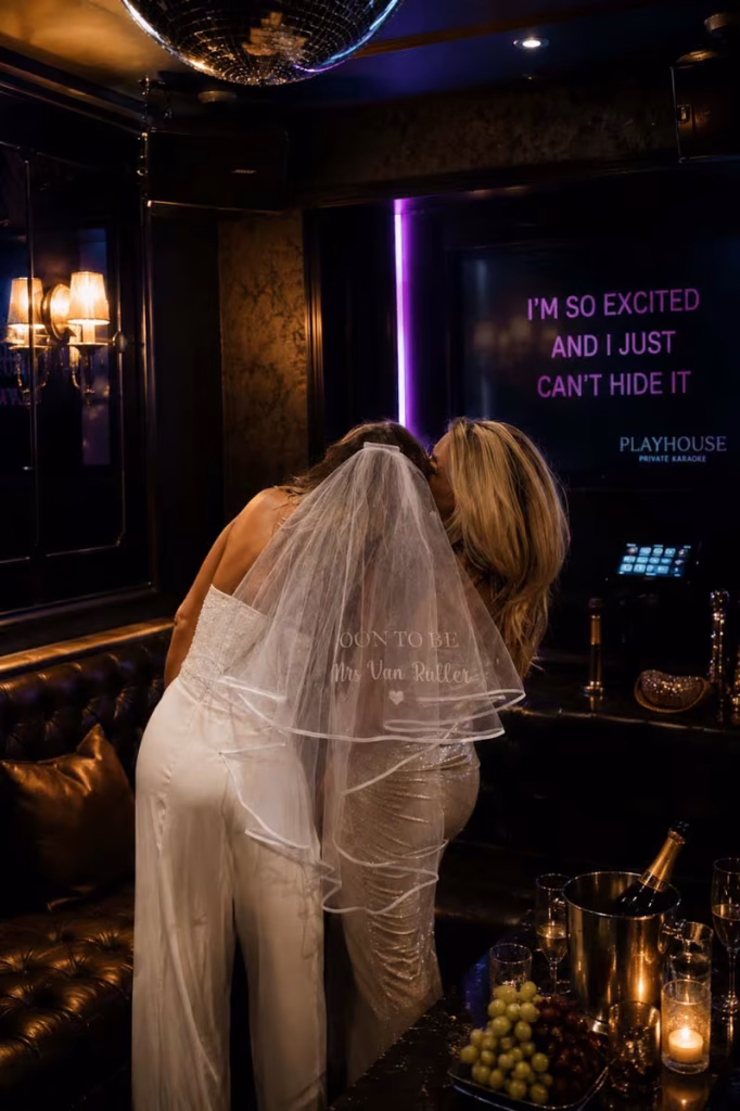 Bride in white dress and personalized veil kissing friend in private karaoke room at Playhouse Paris, champagne and candles on table, neon lyrics on screen in background