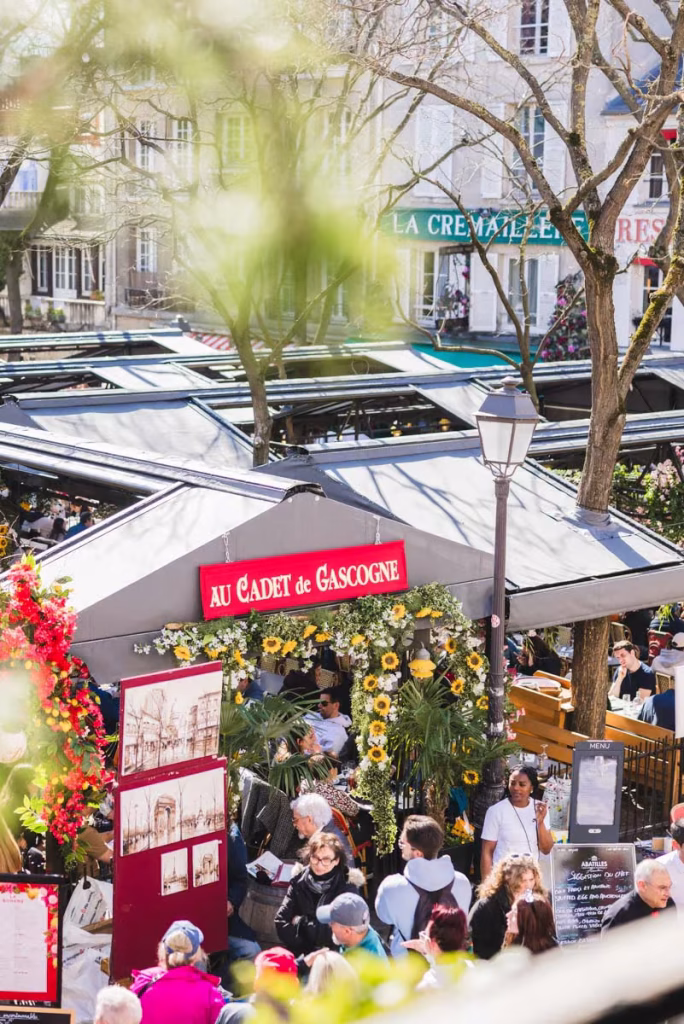 Outdoor terrace of Au Cadet de Gascogne restaurant on Place du Tertre in Montmartre, Paris, decorated with sunflowers and flowers, busy lunch crowd on sunny day