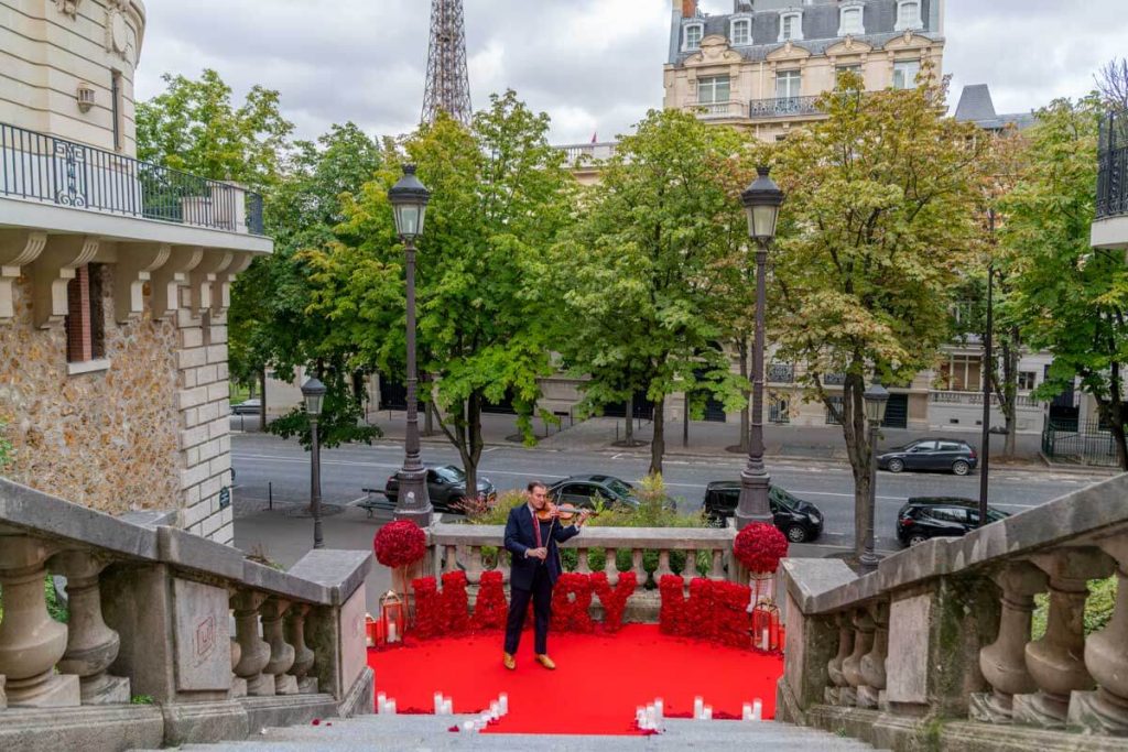 Mise en scène luxueuse pour une demande en mariage sur l'avenue de Camoëns à Paris, avec des lettres rouges formant « Marry Me », un violoniste, un tapis rouge, des bougies et une vue sur la Tour Eiffel.
