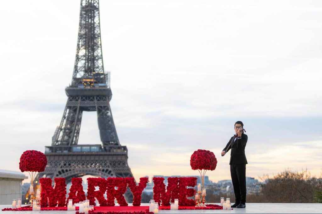Montaje para una propuesta de matrimonio con las letras «Marry Me» en el Trocadéro de París: grandes letras de rosas rojas sobre una alfombra roja con velas, dos topiarios en forma de bola de rosas, un violinista vestido con traje negro tocando junto a la decoración y la Torre Eiffel justo detrás al amanecer.